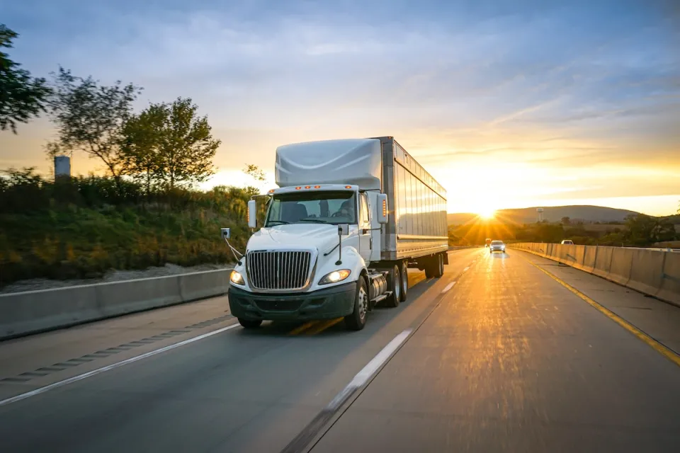 White freight truck on asphalt highway with sunset in background.
